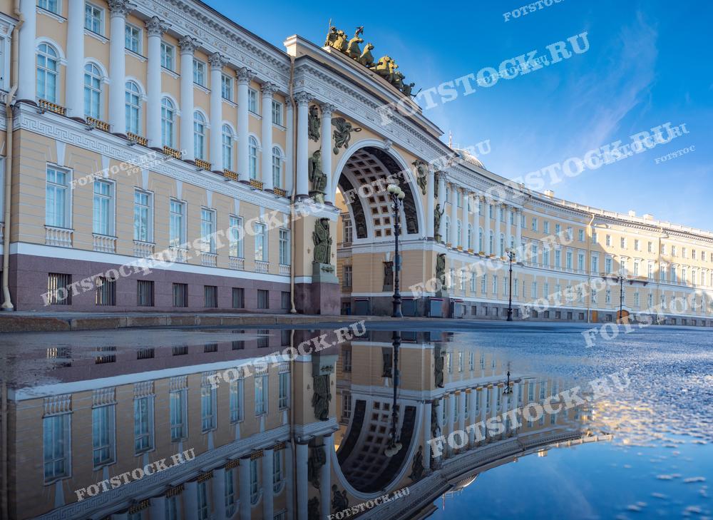 На фотографии изображено здание Главного штаба на Дворцовой площади в Санкт-Петербурге. Утренний свет подчеркивает красоту и величие архитектуры, а отражение здания в луже добавляет снимку глубины и симметрии. Голубое небо и чистый воздух создают ощущение свежести и спокойствия.
